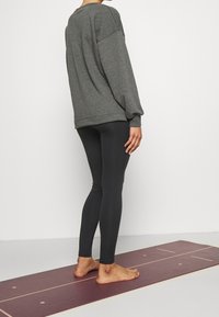 Šedá nadměrná mikina s kleslými rameny, spárovaná s černými legínami. Modelka stojí na vínovém jógovém podložce, bosa, čelí bokem.