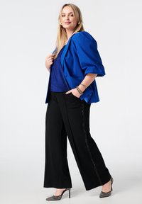 Blauwe blazer met pofmouwen over een glinsterende blauwe top, gecombineerd met zwarte wijde broekspijpen met paillettenaccenten en schoenen met hak.