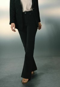 Jeans negre, evazate, dintr-un material moale, asociate cu un top deschis la culoare și un blazer negru, având un design elegant și un minim de accesorii.