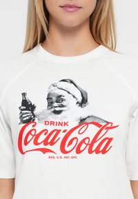 Mladý človek oblečený v bielom tričku s vintage Santa Clausom, ktorý drží fľašu Coca-Coly, a červeným logom "Drink Coca-Cola" naprieč hrudníkom.