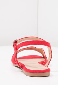 Sandale en daim rouge avec un talon ouvert, deux brides de cheville réglables et une semelle plate beige. Intérieur lisse avec des accents de couture minimalistes.