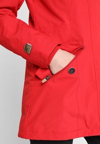 Chaqueta impermeable roja con un bolsillo con cremallera, puños elásticos y un pequeño parche de la marca en la manga. Textura suave y mate.