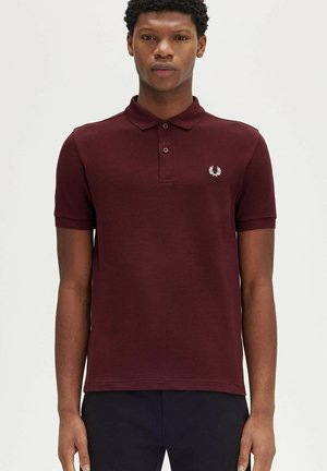 Joven de pie de frente vistiendo una camisa polo burdeos de manga corta con un pequeño logo de laurel blanco en el pecho.