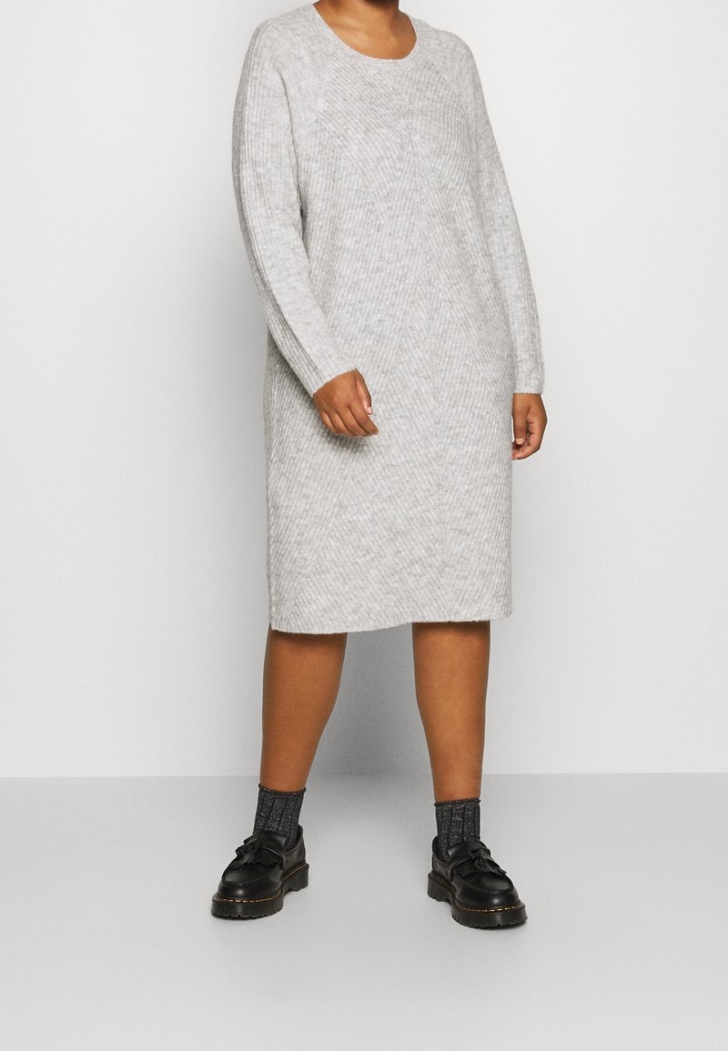 Robe tricot grise avec un col rond, manches longues et texture côtelée. Longueur genou, arborant un motif en diamant subtil. Chaussures noires et chaussettes à motifs.