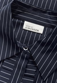 Navyblauwe blouse met witte verticale strepen, met een knoopsluiting aan de voorkant, puntige kraag en een merklabel dat aan de binnenkant is gestikt.