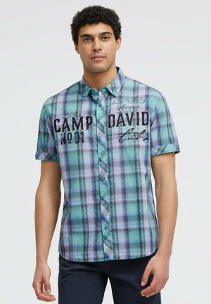 Mann trägt ein kurzärmliges blau-grünes kariertes Hemd mit Knöpfen und dem Text "Camp David" vorne, steht vor einem einfarbigen Hintergrund.
