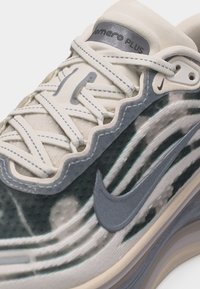 Κοντινό πλάνο λευκού και γκρι παπουτσιού Nike Air Max με ύφασμα με κυματιστά σχέδια, γκρι λογότυπο Swoosh και την επιγραφή "Romero Plus" στην ταμπέλα της γλώσσας.