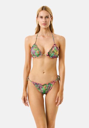 Donna snella che indossa un bikini colorato con motivo floreale e bordi smerlati, in piedi contro uno sfondo bianco semplice.