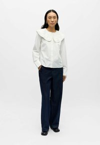 Camisa blanca abotonada con un gran cuello de encaje texturizado, combinada con jeans de mezclilla oscuros de pierna ancha. Zapatos negros brillantes completan el conjunto.