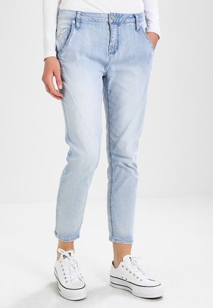 Hellblaue, verjüngte Jeans mit schrägen vorderen Nähten, getragen mit weißem Langarmshirt und weißen Sneakers mit niedrigem Schaft vor neutralem Hintergrund.