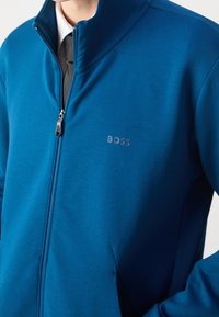 Blå zip-up jacka i slät tyg, med hög krage, sidofickor och en diskret präglad logotyp på bröstet.