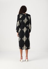 Vestido negro con un elaborado patrón crema, de mangas largas y cintura ceñida con cinturón. La tela parece suave con un dobladillo a la altura de la rodilla. Tacones blancos con tiras visibles.