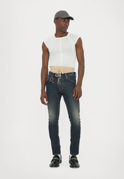 Fitted white sleeveless top, blue denim jeans with light fading, beige drawstring waistband, black loafers, and a gray cap.