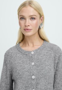 Sivá pletená kardigán so zaobleným výstrihom, ktorý má päť dekoratívnych kvetinových gombíkov. Model má na sebe veľké strieborné geometrické náušnice.