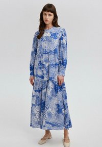 Sinine ja valge pikkade varrukatega kleit lille- ja abstraktses mustris, mitme tasandiga seelikuosa, ümmargune kaelus ja lõdvestunud istuvus. Lamavad, heledad kingad.