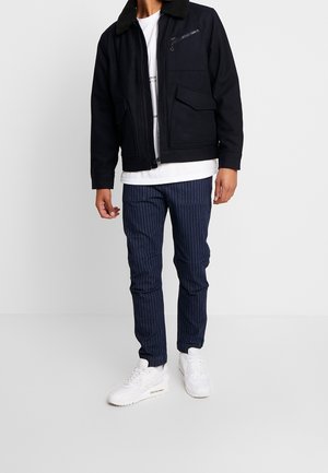 Hombre con chaqueta negra con bolsillos con cremallera, camiseta blanca, pantalones azul marino con rayas finas y zapatillas blancas, posando contra un fondo blanco.