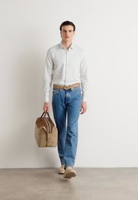 Beige Reisetasche mit braunen Akzenten, gehalten von einer Person in einem hellgrauen Hemd und blauen Jeans, die auf einem strukturierten Boden steht.