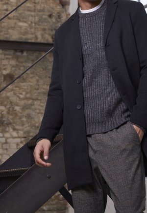 Homme portant un manteau sombre, un pull en maille gris et un pantalon à carreaux, debout à l’intérieur près d’un escalier en métal et d’un mur en pierre.