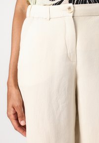 Pantaloni in lino beige con una texture liscia, dotati di chiusura anteriore con bottone, tasche laterali e design a gamba dritta.