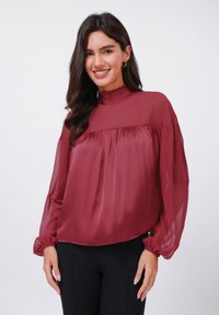 Bordeauxfarbige Bluse mit hohem Kragen, langen durchsichtigen Ärmeln und gerafftem Oberteil. Weicher Stoff mit fließendem Design und elastischen Bündchen.