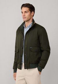 Uomo che indossa una giacca bomber verde scuro sopra una camicia a righe blu con pantaloni beige, in piedi di fronte a uno sfondo chiaro e uniforme.