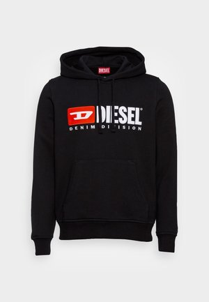 Person trägt zerrissene, weite Jeans, einen schwarzen Diesel-Hoodie, einen grauen Blazer und schwarze Schuhe und steht auf einem gepflasterten Gehweg vor einer Betonwand.