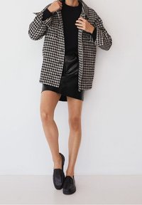 Mustavalkoinen houndstooth-kuvioinen ylisuuri takki, musta teksturoitu lyhyet hame ja mustat loaferit. Sujuvat tekstuurit ja kerrokselliset muotoiluelementit näkyvissä.