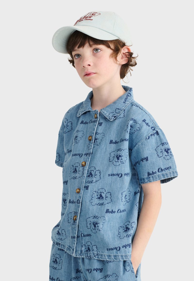 Enfant portant une casquette bleu clair et une chemise à manches courtes en denim assortie ainsi que des shorts avec des motifs de têtes de chien cartoon et le texte "Bobo Choses".
