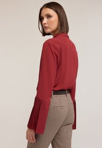 Blouse rouge à manches longues plissées, associée à un pantalon à carreaux taille haute avec une ceinture noire. Tissu lisse, design structuré.