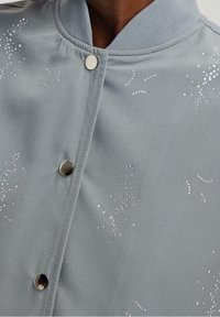 Lys grå bomberjakke med ribbet krave, prydet med metalliske trykknapper og pynt af klare rhinsten langs den ene side.