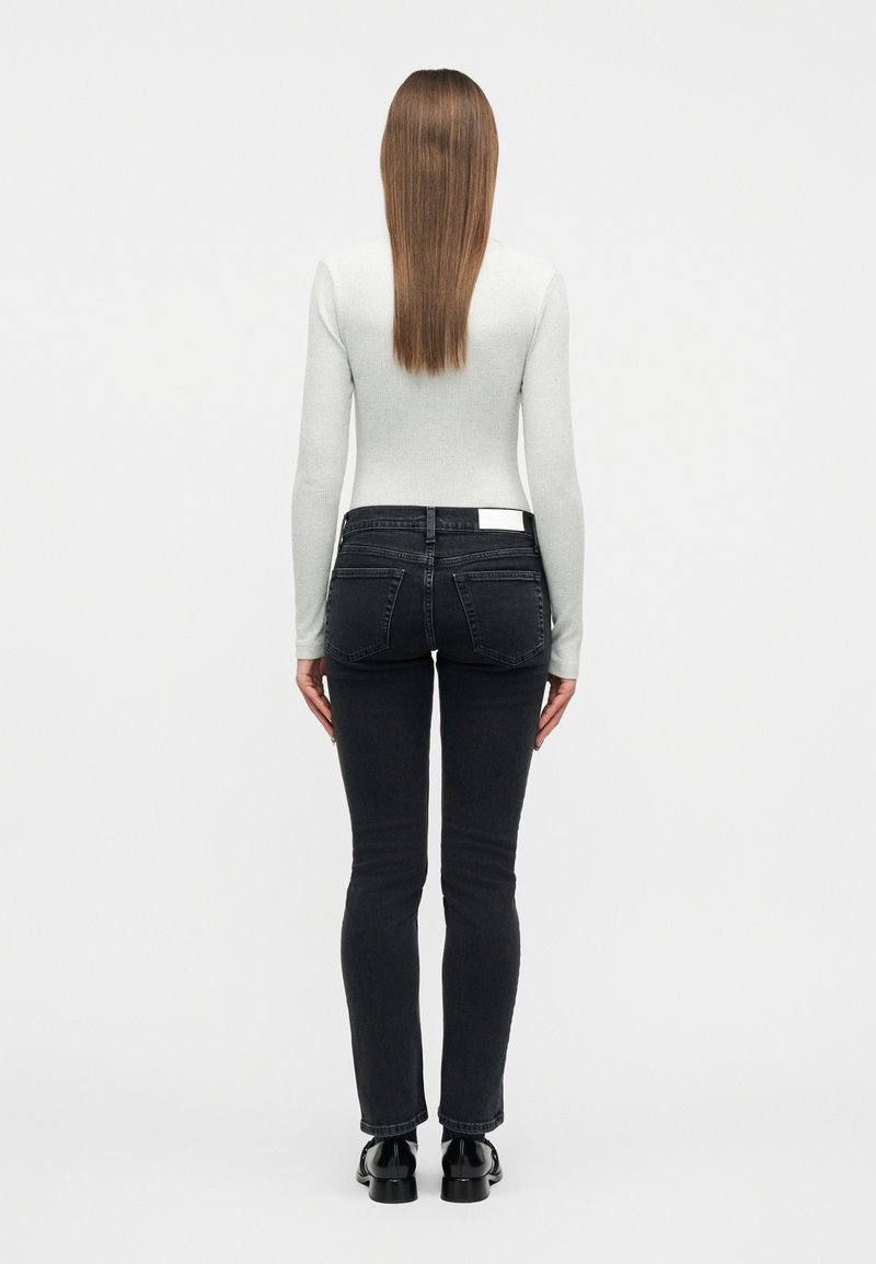 Woman with straight brown hair wearing a light gray long-sleeve top, black slim-fit jeans, and black shoes standing with back facing forward.