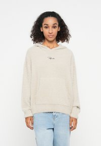 Beige fleece hoodie met een zachte textuur, voorzien van een voorkantzak en een capuchon. Zwart geborduurd logo op de borst. Draagt samen met lichtblauwe jeans.