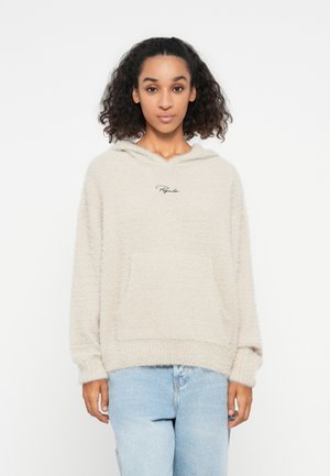 Beige Fleece-Hoodie mit weicher Textur, ausgestattet mit einer Fronttasche und einer Kapuze. Schwarze Logo-Stickerei auf der Brust. Getragen mit hellblauen Jeans.
