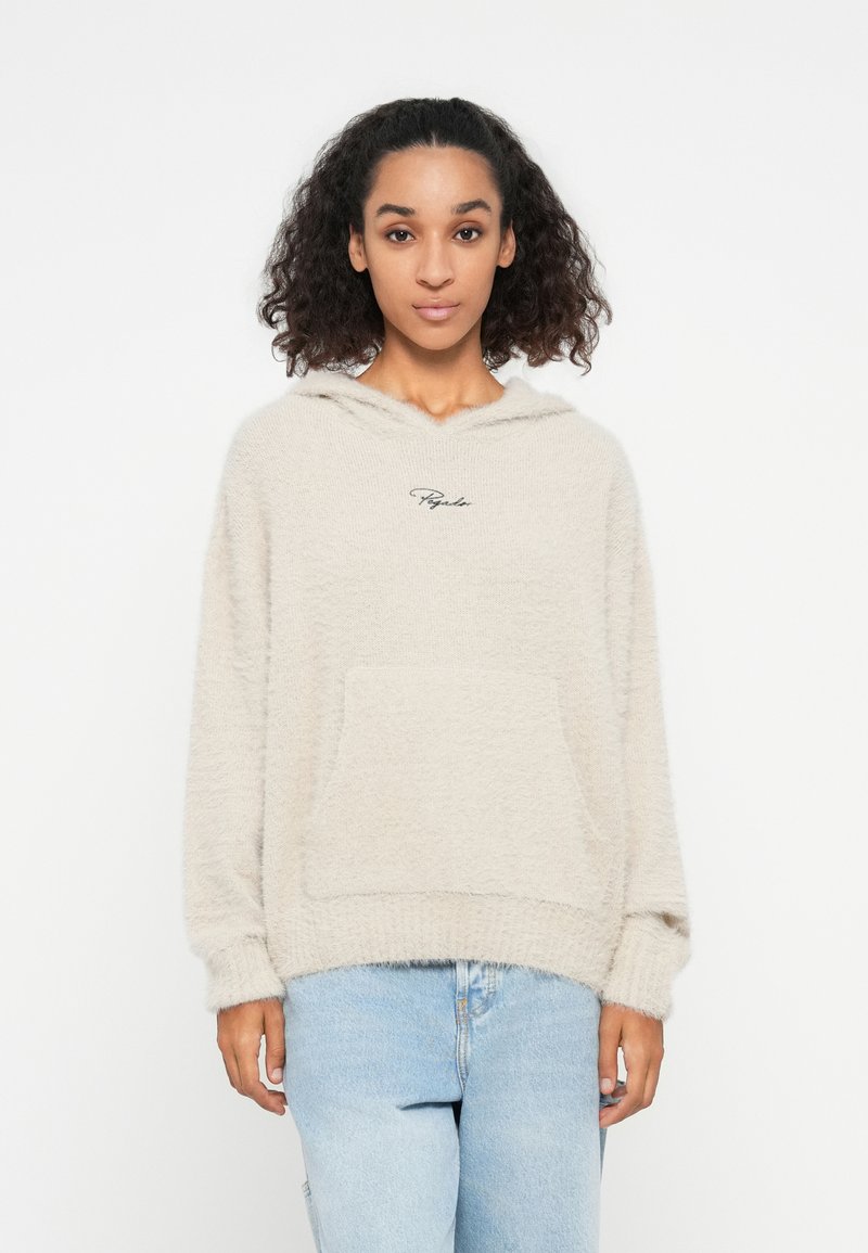 Beige fleece hoodie met een zachte textuur, voorzien van een voorkantzak en een capuchon. Zwart geborduurd logo op de borst. Draagt samen met lichtblauwe jeans.