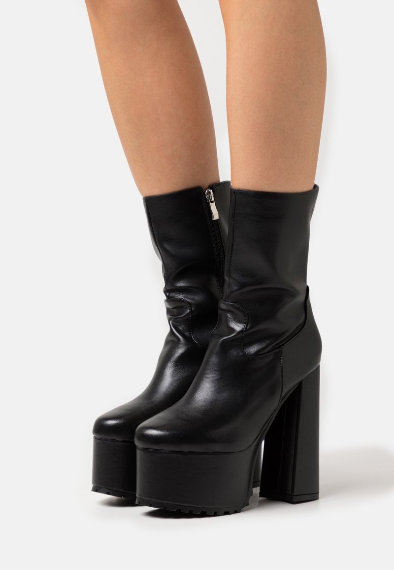 Bottines à plateforme en cuir noir avec talons hauts épais et fermeture éclair latérale, portées sur des jambes nues sur fond blanc.