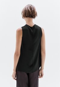 Top nero senza maniche con scollatura rotonda, dettagli di cucitura sottili, orlo leggermente curvato, indossato con pantaloni scuri larghi.