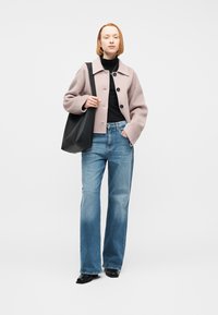 Γυναίκα με κόκκινα μαλλιά που φοράει ελαφρύ μωβ σακάκι, μαύρο ζιβάγκο, μπλε τζιν, μαύρα παπούτσια και κρατά μια μεγάλη μαύρη τσάντα ώμου.