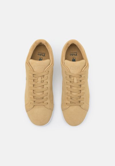Sapatilhas de camurça em tom claro com solas de borracha, apresentando um design com atacadores, bico redondo e um logótipo subtil na lingueta. Textura suave, estilo minimalista.