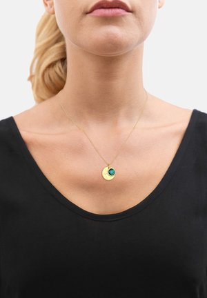 Collana d'oro con un pendente circolare texture e una piccola pietra verde. Indossata su una maglietta nera, evidenziando il suo design e il contrasto di colore.