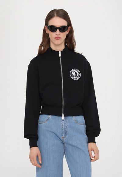 Melnā zipa cropped sporta jaka ar augstu apkakli un priekšējo kabatu, kurai ir balts apaļš logo uzlīme. Modelis valkā melnas saules brilles.
