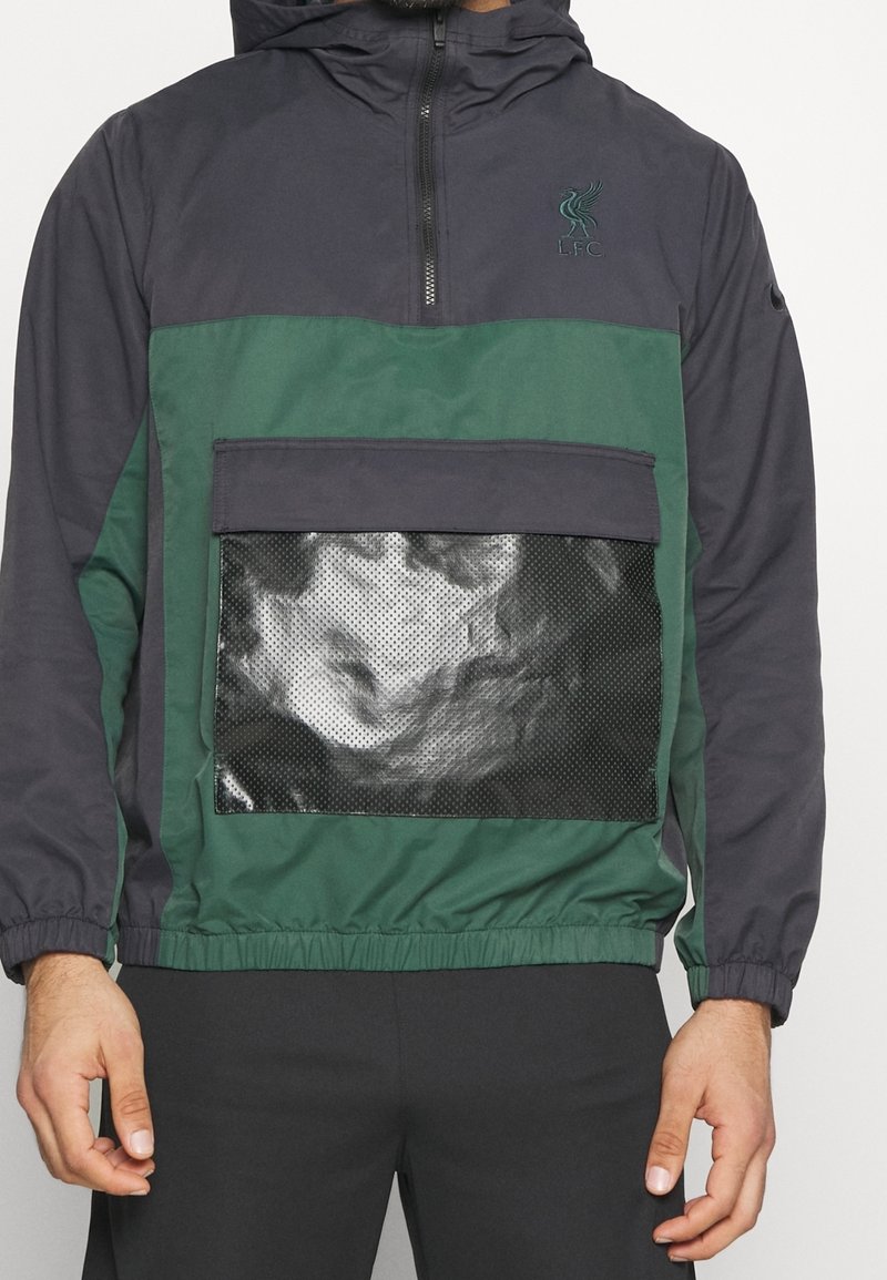 Veste de sport noire et verte à demi-fermeture éclair avec logo du Liverpool FC, poche avant en maille, poignets et ceinture élastiques, portée par une personne.