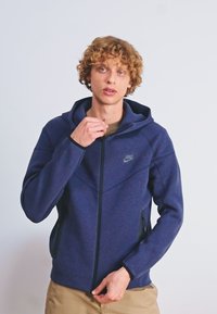 Felpa con zip di colore blu navy, realizzata in un tessuto testurizzato, con tasche anteriori e cappuccio regolabile con cordino. Presenta un piccolo logo sul lato sinistro superiore.