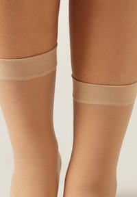 Nude strømper til låret med en glat, gennemsigtig tekstur og lys beige farve. Bløde, elastiske bånd øverst for en sikker pasform.