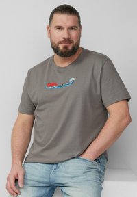 Grijze katoenen t-shirt met rode "JAWS" tekst en een golfontwerp met een haai, gecombineerd met lichtblauwe denim jeans.