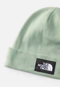 Berretto lavorato a maglia verde menta con risvolto e un'etichetta nera con testo e logo bianchi. Texture liscia, vestibilità elastica.