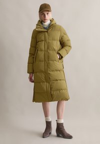 Olijf groene pufferjas met een middellange snit, ritssluiting, zijzakken en geribde manchet. Gecombineerd met bruin met hakken en een pet.