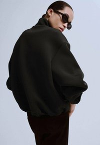 Czarna, oversize'owa bluza z wysokim kołnierzem, opuszczonymi ramionami i fakturowaną tkaniną. Modelka ozdobiona ciemnymi okularami przeciwsłonecznymi.