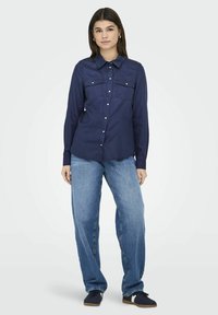 Camicia blu navy con bottoni, due tasche sul petto, maniche lunghe e collo classico, abbinata a jeans a gamba larga azzurri e sneakers scure.