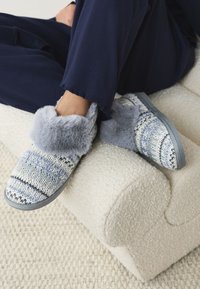 Chaussons tricotés bleus et gris avec un revers en peluche grise. Texture douce, forme arrondie et semelle incurvée. Reposant sur une surface claire texturée.