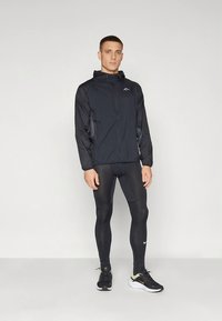 Zwarte sportieve jas met ritssluiting aan de voorkant, voorzien van een capuchon en contrasterende stoffen panelen. Gecombineerd met zwarte leggings en zwarte sneakers.